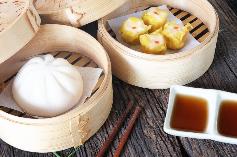 Gedämpfte Dim Sum in einem Bambuskorb mit Soßen und Essstäbchen auf Holzoberfläche.