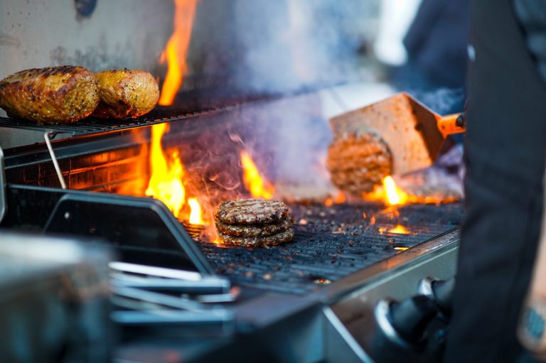 Fleisch-Patties braten auf einem Grill mit lebhaften Flammen im Hintergrund.