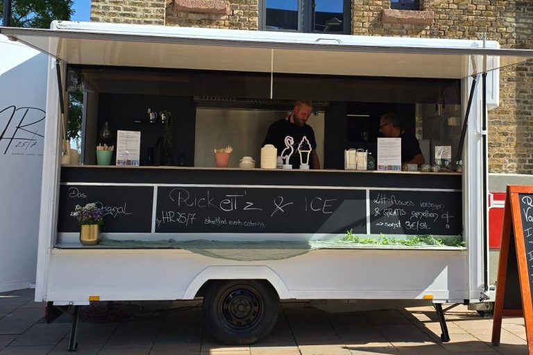 Foodwagen mit offenem Fenster, zwei Personen bereiten Speisen und Getränke vor.