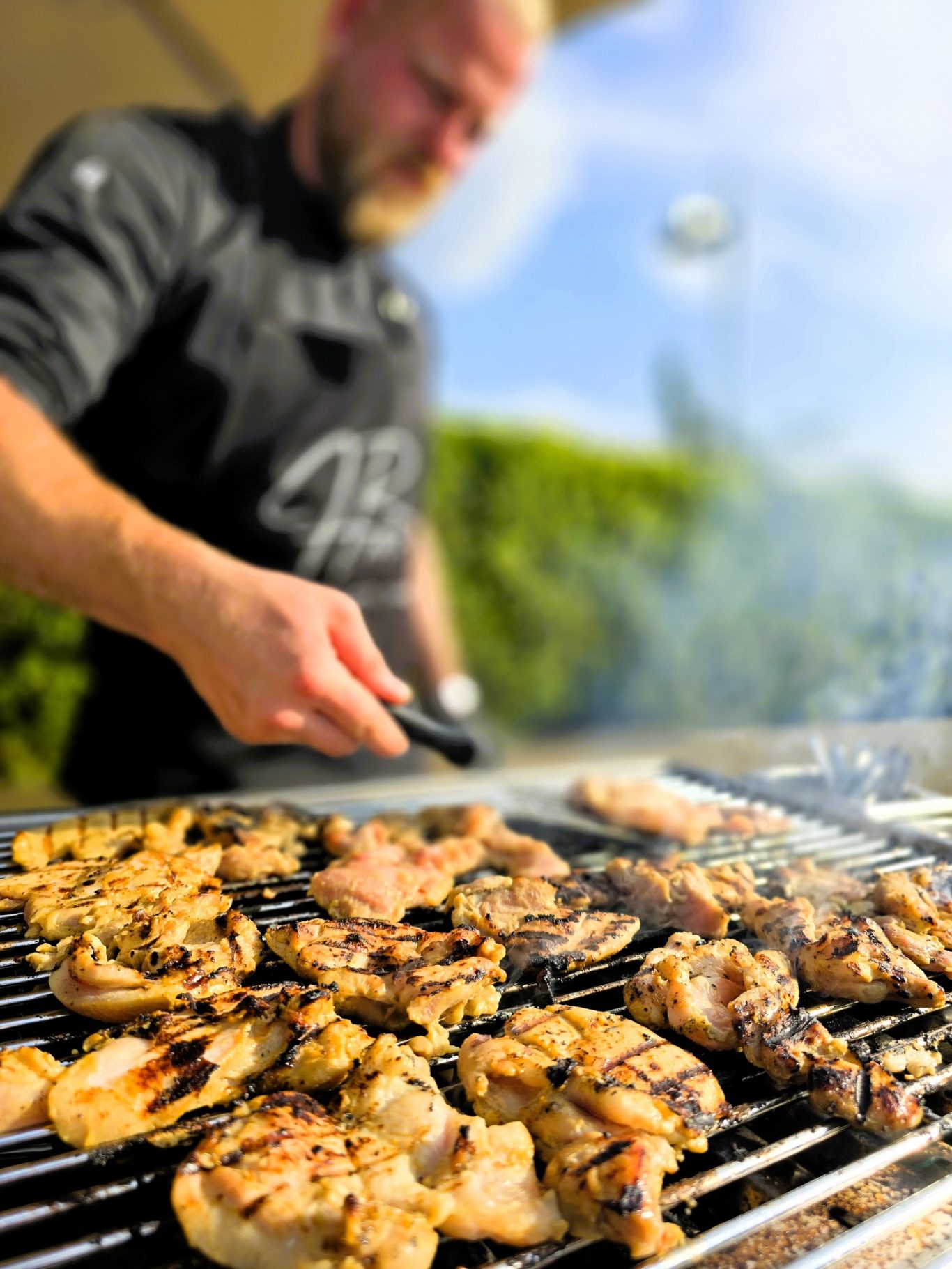 Ein Mann grillt vielfältige Fleischstücke im Freien. Sonne scheint.