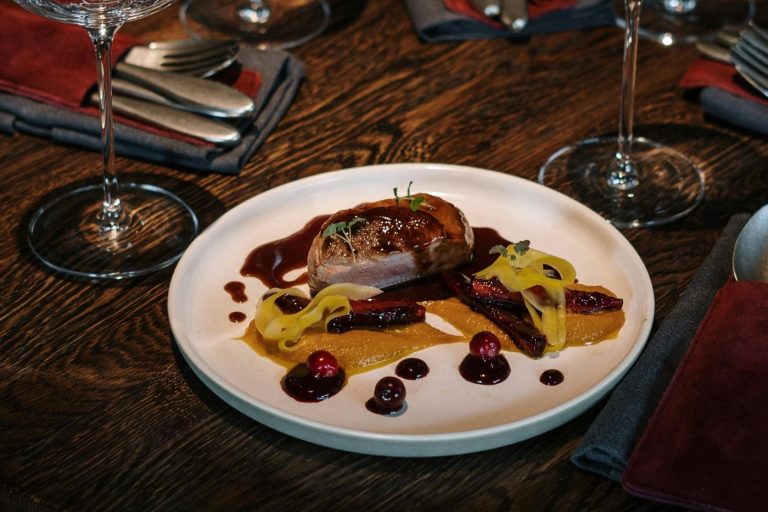 Gericht mit Fleisch, Soße, Püree und Beeren auf einem weißen Teller, umgeben von Gläsern.
