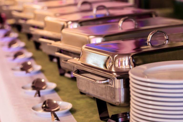 Buffet mit mehreren warmen Speisen in Edelstahlbehältern und Tellern mit Desserts.
